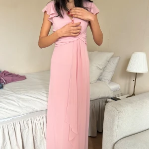 Ljusrosa långklänning från TFNC London - Långklänning i ljusrosa från TFNC London med omlottliv, v-ringning och korta volangärmar. Klänningen har en knytdetalj i midjan och är tillverkad i ett lätt, flytande material som ger ett elegant fall. Dragkedja bak för enkel påklädning.