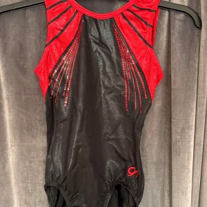 Svart och röd gymnastikdräkt GK - Säljer en snygg gymnastikdräkt från GK Elite i svart och rött med glittriga röda detaljer och strass framtill. Dräkten har ärmlös design, hög halsringning och coola band i ryggen. Perfekt för träning eller tävling inom gymnastik.