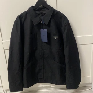 Prada vind/höst jacka  - Svart vind/höst jacka med klassisk krage och diskret logga på bröstet. Jackan har dragkedja framtill, två stora fickor och en stilren, minimalistisk look. Perfekt för dig som gillar exklusiva och cleana plagg.