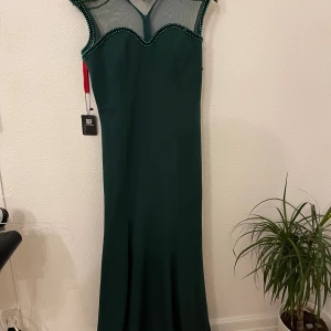 Mörkgrön långklänning med pärldetaljer - Snygg mörkgrön långklänning från Maya Deluxe med mesh upptill och pärldetaljer längs halsringningen och axlarna. Klänningen har en figurnära passform och är ärmlös med en elegant, vågig kant upptill. Perfekt för dig som vill sticka ut med coola detaljer.