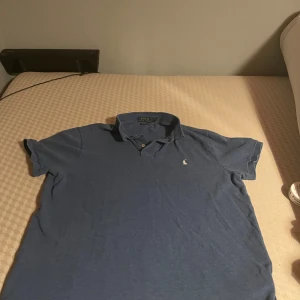 Blå pikétröja från Polo Ralph Lauren - Snygg blå pikétröja från Polo Ralph Lauren i custom slim fit. Tröjan har klassisk krage, korta ärmar och den ikoniska broderade polospelaren på bröstet. Perfekt för dig som gillar stilrena och tidlösa plagg med en sportig touch.