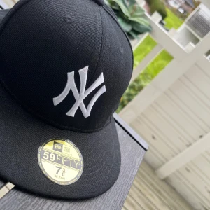 Svart New Era 59FIFTY NY Yankees keps - Snygg svart New Era 59FIFTY keps med klassiskt New York Yankees broderat i vitt framtill och MLB-logga baktill. Platt skärm och stilren look, perfekt för dig som vill ha streetstyle. Tillverkad i bomull och polyester för skön passform. Storlek 58,7 cm