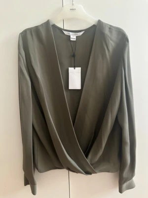 Olivgrön omlottblus från Diane von Furstenberg - Snygg olivgrön omlottblus från Diane von Furstenberg med lång ärm och breda manschetter. Blusen har en djup v-ringning och ett elegant omlottfall framtill. Materialet är mjukt och har en lätt glans, vilket ger en lyxig känsla. Perfekt för dig som gillar stilrena och trendiga plagg. Blusen är i storlek US 6