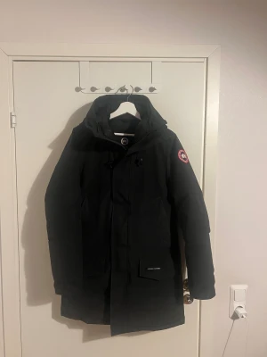 Svart dunjacka från Canada Goose Langford Parka - Säljer en svart dunjacka från Canada Goose med huva och klassisk logga på ärmen. Jackan har långa ärmar, stora fickor framtill och stängs med dragkedja och knappar. Perfekt för kalla vinterdagar och riktigt snygg streetstil.