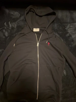 AMI PARIS full zip svart - Svart hoodie från Ami med huva och dragkedja framtill. Tröjan har ett rött broderat hjärta med bokstaven A på bröstet, samt praktiska fickor och snörning vid huvan. Materialet är mjuk bomull och passformen är normal.