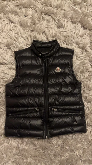 Svart dunväst från Moncler - Snygg svart dunväst från Moncler med quiltad design och hög krage. Västen har två dragkedjeförsedda fickor framtill och ett broderat Moncler-märke på bröstet. Materialet är glansigt och känns lätt men ändå värmande. Perfekt att slänga över en hoodie för en chill look. Säljer då jag är sugen på något nytt. Finns såklart också qr kod att skanna. Ända nackdelen är att det är en liten skråma under vänster ficka som ni ser på bild 7 men det är absolut inget man märker av