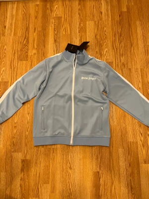 Ljusblå tröja från Palm Angels - Palm Angels ljusblå tröja med vita ränder längs ärmarna och vit dragkedja framtill. Tröjan har ståkrage, två fickor med dragkedja och logotyp på bröstet. Perfekt för dig som gillar streetwear.