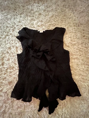 Svart blus med volang från Gina Tricot - Säljer en svart blus från Gina Tricot i storlek 146/152. Blusen har snygga volangdetaljer framtill och längs nederkanten, samt ett coolt strukturmönster i tyget. Ärmlös modell som ger en trendig look och passar perfekt till jeans eller kjol.