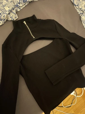 Svart ribbad bolerotopp med dragkedja - Säljer en svart bolerotopp med ribbad struktur och lång ärm. Toppen har en hög krage med silverfärgad dragkedja och en öppen ryggdetalj som ger en edgy vibe. Perfekt för dig som gillar att sticka ut med din stil.