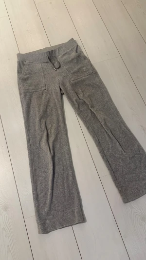 Grå mjukisbyxor Juicy Couture S - Grå mjukisbyxor från Juicy Couture med broderad logga på fickan, snörning i midjan och bakfickor med blingiga knappar. Sköna och loose fit med raka ben, tillverkade i mjukt bomullsmaterial. Perfekta för chill dagar. Nypris 1200kr
