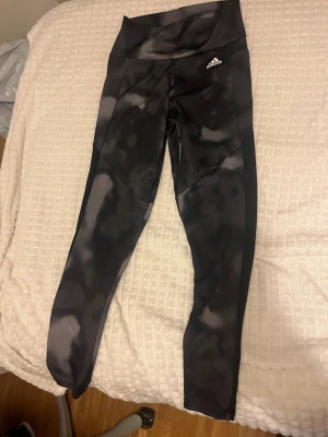 Svarta Adidas leggings med mönster - Snygga svarta leggings från Adidas med coolt mönster i grått och svart. Hög midja och smal passform som sitter skönt under träning eller chill. Adidas-logga på höften. Materialet är stretchigt och känns mjukt mot huden.