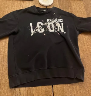 Svart Dsquared2 ICON sweatshirt L - Svart sweatshirt från Dsquared2 i storlek L med stort vitt 'ICON'-tryck och färgstänk på bröstet. Tröjan har rund halsringning och är tillverkad i mjuk bomull. Perfekt för dig som vill sticka ut med en cool streetstyle-look.