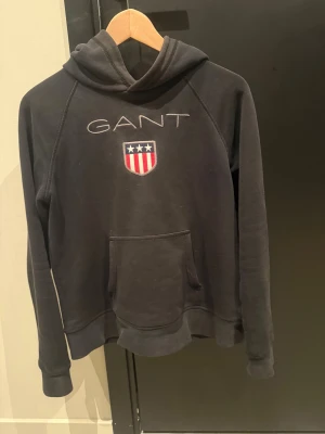 Svart hoodie från GANT med emblem - Svart hoodie från GANT med broderat emblem och logga på bröstet. Tröjan har huva, lång ärm och en stor magficka framtill. Materialet är mjuk bomull och polyester, perfekt för chill dagar. Klassisk och stilren design med amerikansk touch.