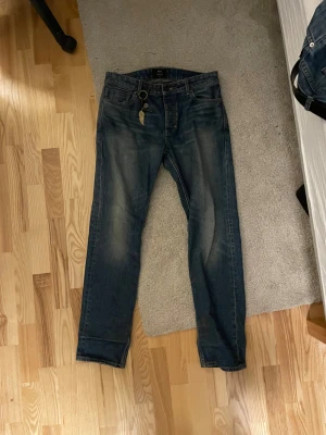 Blå jeans från Neuw Stockholm - Säljer ett par snygga blå jeans från Neuw Stockholm med klassisk femficksdesign och coola slitningar på låren. Jeansen har en rak passform och är tillverkade i robust denim med detaljerade sömmar. Perfekt för dig som gillar stilrena och tidlösa jeans. Nypris 1600