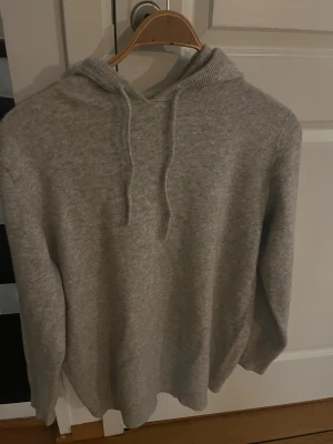 Grå stickad hoodie med snörning - Mysig grå merinoull hoodie med stickad känsla och snörning i huvan. Tröjan har långa ärmar och ribbad kant vid nederdelen och ärmsluten. Perfekt för chill dagar och när du vill ha det extra bekvämt.