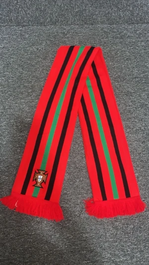 Portugal halsduk med ränder och emblem - Snygg halsduk i rött med svarta och gröna ränder, officiellt Portugal-emblem broderat på båda sidor. Långa fransar i ändarna ger extra stil. Perfekt för att visa ditt stöd på matchen! Materialet är 100% akryl och halsduken är producerad i Portugal.