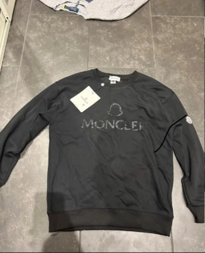 Svart sweatshirt från Moncler - Svart sweatshirt från Moncler med diskret logotyp i svart på bröstet och klassisk Moncler-patch på ärmen. Tröjan har rund hals, långa ärmar och ribbade muddar. Perfekt för dig som gillar stilrena och exklusiva märken.