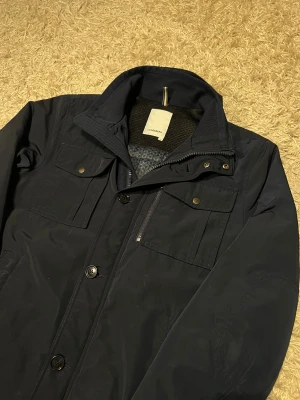 J.Lindeberg fieldjacket trenchcoat herr M svart marinblå - J.Lindeberg fieldjacket i svart/marinblå färg. Storlek M med justerbar rygg för passform. Funkar bra till både höst och vinter. Mycket bra skick, har lite små defekter inget som märks, se bild. Postas antigen samma dag som köp eller dagen därpå🙌💯