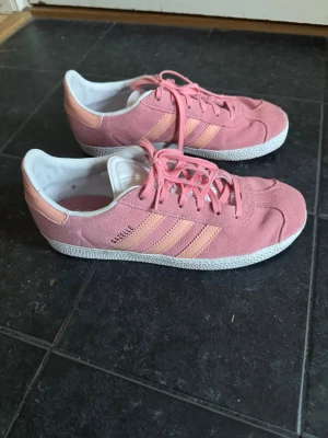 Adidas Gazelle rosa sneakers mycket bra skick  - Säljer ett par snygga Adidas Gazelle sneakers i rosa mocka med klassiska tre ränder i ljusrosa på sidorna. Skorna har vit sula, snörning och rund tå. Perfekta för dig som vill ha en färgklick till outfiten. Endast använda ca 10 gånger. Storlek 37 1/3