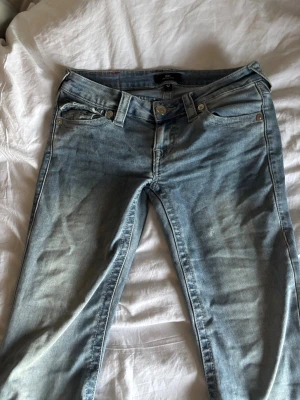 True Religion Joey Low Rise Flare Jeans - Snygga ljusblå jeans från True Religion, modell Joey Low Rise Flare. De har låg midja, utsvängda ben och klassiska fem fickor. Jeansen är i bomull och har slitna detaljer vid bensluten för en cool, avslappnad look.