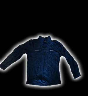 Mörkblå henley från Patagonia - Mörkblå långärmad henley från Patagonia med halv dragkedja framtill och ståkrage. Skjortan har en clean look med diskret logga på bröstet och är tillverkad i lätt polyester som andas. Perfekt för dig som gillar outdoor och sportig stil.