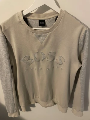 Hugo Boss tröja - Snygg beige och grå sweatshirt från Hugo Boss med broderad logga på bröstet. Tröjan har ribbad rund hals, muddar och långärmade gråmelerade ärmar. Perfekt för chill dagar och enkel att matcha med jeans eller joggers.