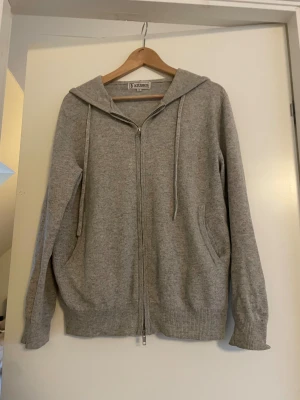 Grå hoodie från Studios med dragkedja - Säljer en grå hoodie från Studios i storlek M. Tröjan har huva med snörning, hel dragkedja framtill och två fickor på sidorna. Materialet är mjukt och känns som bomull eller syntet, perfekt för chill dagar. Ribbad nederkant och ärmslut ger en snygg finish. Säg till om ni har några funderingar 