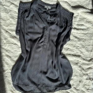 Mörkgrå blus från Noa Noa - Säljer en mörkgrå blus från Noa Noa i mjuk viskos. Blusen har korta ärmar, knappar framtill och snygga smockdetaljer på axlarna. Den har en avslappnad passform och linneliknande tyg som ger en stilren look. Storlek markerad 36, men passar även 38. Se mått