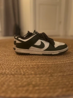 Nike Dunk Low i vitt och mörkgrönt - Nike Dunk Low sneakers med vit bas och mörkgröna paneler. Klassisk låg siluett med rund tå, snörning och platt sula. Swoosh-logga på sidan och vadderad krage för extra komfort. Perfekt för dig som gillar streetwear och sportig stil.