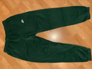 Gröna mjukisbyxor från Nike - Mörkgröna mjukisbyxor från Nike med klassisk logga i vitt på vänster lår. Byxorna har elastisk midja och muddar vid bensluten för en skön och avslappnad passform. Perfekta för chill eller träning.
