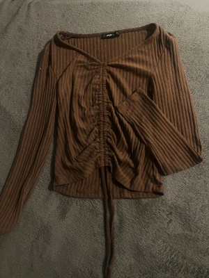 Brun ribbad omlottopp med snörning - Säljer en brun långärmad topp med ribbad struktur och snygg snörning framtill. Toppen har omlott-look och v-ringad hals.