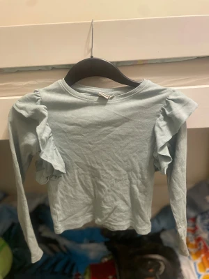 Ljusblå ribbad topp med volang SRN BILT - Supersöt ljusblå långarmad topp från SRN BILT i ribbad bomull. Toppen har volangdetaljer på axlarna som ger en extra touch och en rund halsringning. Perfekt för dig som gillar stilrena och mjuka plagg. Barn till tjejer, storlek 134/140