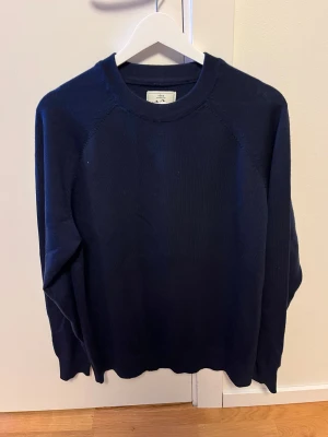 Marinblå merinoulltröja från Yoke - Säljer en stilren marinblå tröja från Yoke i 100% merinoull. Tröjan har rund hals, lång ärm och är stickad med mjuk känsla. Perfekt för dig som gillar klassisk look och vill ha något som är både bekvämt och snyggt.