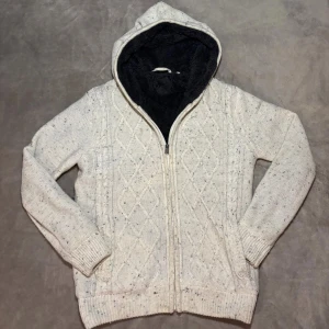 Fodrad Cardigan - Mysig vit stickad hoodie med dragkedja och huva fodrad i svart fleece. Tröjan har ett klassiskt kabelstickat mönster framtill och ribbade muddar. Perfekt för kyliga dagar när du vill vara både varm och snygg.