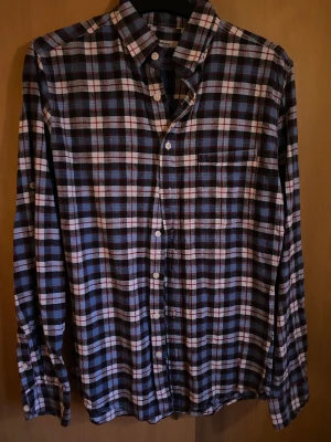 Rutig blå och vit skjorta - Säljer en klassisk rutig skjorta i blått, vitt och rött med lång ärm och knappar framtill. Skjortan har en bröstficka och en snygg krage som ger en chill och stilren vibe. Perfekt för dig som gillar avslappnad men ändå schysst stil.