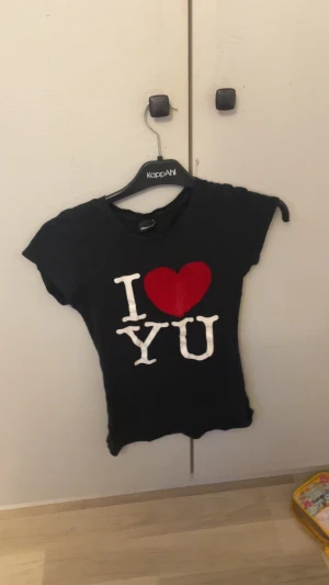 Svart t-shirt med I ❤️ YOU-tryck - Svart t-shirt från KappAhl med korta ärmar och ett stort tryck framtill med texten 'I ❤️ YOU' i vitt och rött. T-shirten har en klassisk rund halsringning och är gjord i mjuk bomull. Perfekt för dig som vill ha en enkel men uttrycksfull look.