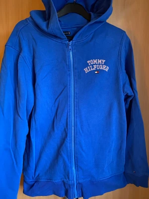 Blå hoodiejacka från Tommy Hilfiger - Blå hoodiejacka från Tommy Hilfiger med dragkedja framtill och logotyp på bröstet. Jackan har huva, ribbade muddar och är långärmad. Perfekt för chill dagar och enkel att matcha med jeans eller joggers.