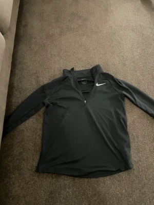 Svart Nike half zip Dri-Fit tröja XL 158-164 - Svart långärmad tröja från Nike i storlek XL, med vit logga på bröstet och half zip-dragkedja. Tillverkad i Dri-Fit polyester som andas och är perfekt för träning. Snygg och sportig passform med hög krage.