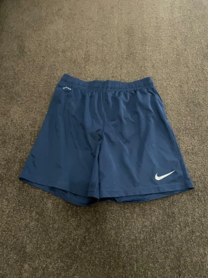 Blå Dri-Fit shorts från Nike - Säljer ett par blå Nike Dri-Fit shorts med elastisk midja och klassisk vit swoosh-logga broderad på benet. Perfekta för träning eller sport, tillverkade i lätt och ventilerande syntetmaterial som håller dig fräsch. Enkel design med loose passform.