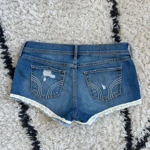 Blå shorts med spets från Hollister - Snygga blå jeansshorts från Hollister i storlek w29, short short med låg midja, slitna detaljer och råa kanter. Shortsen har vit spets längs bensluten och klassiska fickor både fram och bak. Midjemått 43cm mätt rakt över 💙