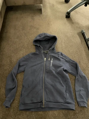 Mörkblå hoodie från Sail Racing - Mörkblå hoodie från Sail Racing med hel dragkedja och extra zip-ficka på bröstet. Snyggt broderad logga på framsidan och huva med snörning. Tillverkad i mjuk bomull, perfekt för chill eller häng med kompisar. Långärmad och normal passform.