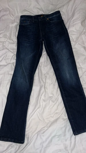 Mörkblå jeans från BOSS - Snygga mörkblå jeans från BOSS med klassisk femficksdesign och subtila slitningar. Jeansen har raka ben och orangea kontrastsömmar, samt BOSS-logga på både knapp och ficka. Perfekta för en stilren och avslappnad look.