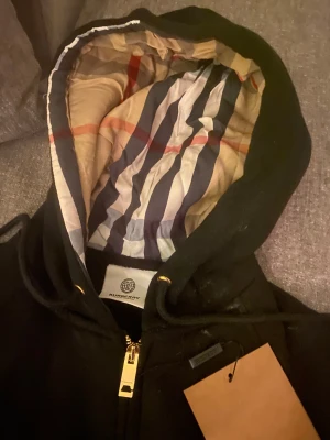 Svart Burberry hoodie med dragkedja - Svart hoodie från Burberry med klassisk rutigt foder i huvan och guldfärgad dragkedja. Tröjan har snörning vid huvan och ribbade muddar. Perfekt för dig som vill ha en stilren och lyxig look med ikoniska Burberry-detaljer.