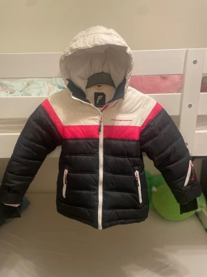 Pufferjacka från Five Seasons, 134/140 - Snygg pufferjacka från Five Seasons i färgerna svart, vit och rosa. Jackan har huva, flera dragkedjor med rosa detaljer och broderad logga på bröstet och armen. Tillverkad i polyester och har quiltad design som håller värmen under kalla dagar. Barn till tjej 