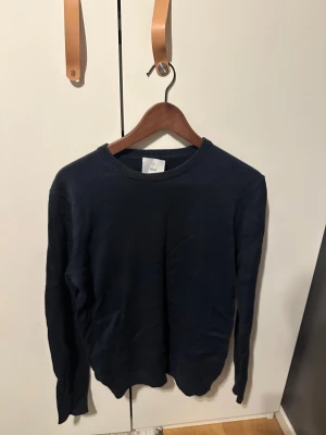 Mörkblå långärmad tröja - En stilren mörkblå tröja från Bläck i klassisk modell med rund hals och ribbade muddar. Tröjan är tillverkad i mjuk bomull och har en enkel, clean look som funkar till jeans eller chinos. Perfekt för dig som gillar basic och tidlöst.