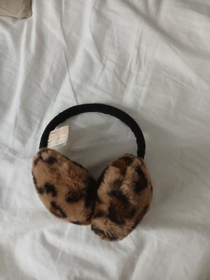 Leopardmönstrade öronmuffar diadem - Snygga öronmuffar med leopardmönster och fluffig pälsimitation. Diademet är svart och mjukt, perfekt för att hålla öronen varma och ge din vinterstil en cool touch. Passar dig som gillar djurmönster och vill sticka ut.