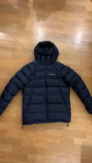 Peak Performance jacka - Säljer en svart dunjacka från Peak Performance med huva och dragkedja framtill. Jackan har en quiltad design, två sidofickor med dragkedja och justerbar nederkant. Perfekt för kalla dagar och riktigt snygg till streetwear-stilen. Storlek M. Mycket bra skick