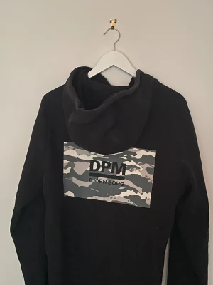 Svart hoodie Björn Borg, storlek L - Svart hoodie från Björn Borg med coolt DPM-camouflageprint på ryggen. Tröjan har huva med snörning och långa ärmar. Materialet är mjukt och skönt, perfekt för chill eller sport. Snygga metalländar på snörena ger extra stilpoäng.