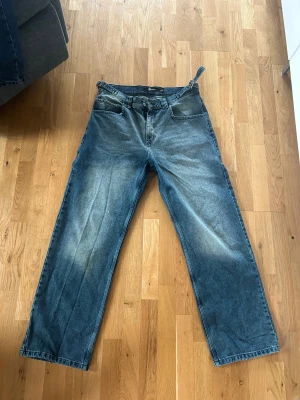 Blåa vida jeans med gröna detaljer - Säljer ett par blåa jeans med bred passform och snygg vintage tvätt med lite gröna detaljer. Jeansen har klassiska fem fickor, raka ben och hög midja. Materialet är robust denim som ger en cool och avslappnad look. Perfekta för dig som gillar loose fit och streetstyle.
