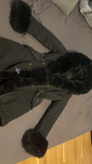 Svart parkajacka med pälsdetaljer - Svart parkajacka från Capsule med fluffiga pälsdetaljer på huva och ärmslut. Jackan har dragkedja och knappar framtill, praktiska fickor och snörning i midjan för snygg passform. Perfekt för kalla dagar med sin långa modell och varma känsla.